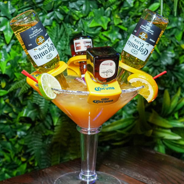 A large margarita glass with orange liquid, garnished with lime, orange, two Coronitas, and two Don Julio mini bottles.